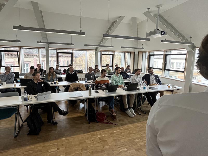 Teilnehmer sitzen in einem hellen Seminarraum mit Laptops und hören einem Vortrag zu.