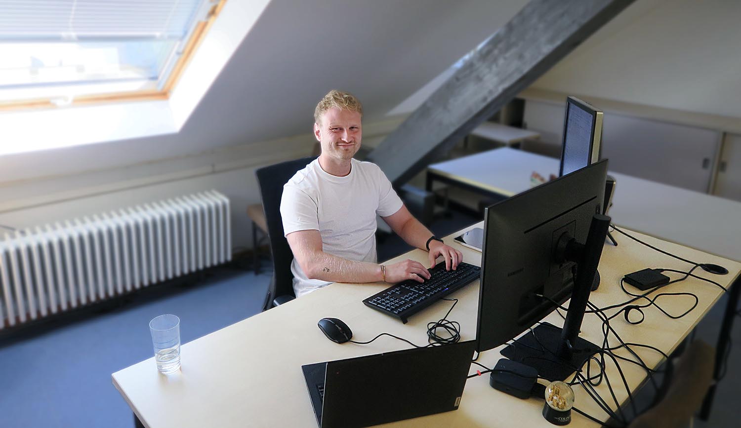 Person sitzt mit Laptop am Schreibtisch und lächelt in die Kamera.