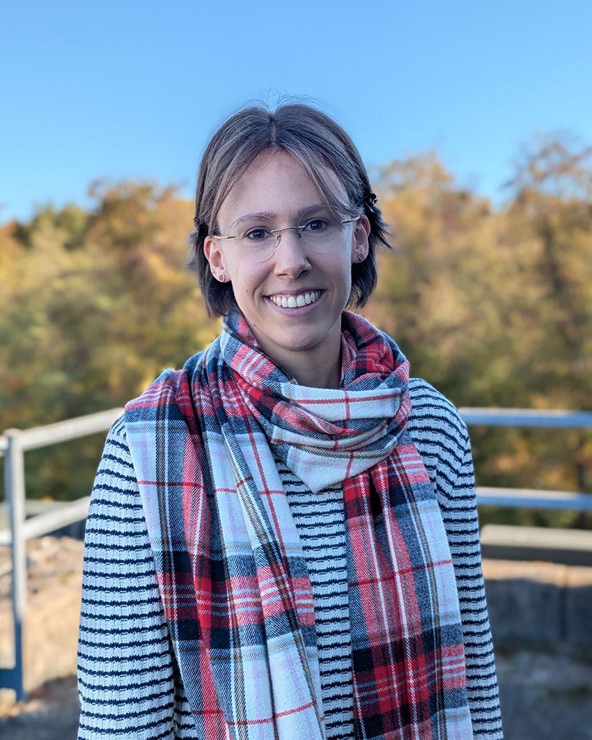 Eine lächelnde Person mit Brille trägt ein gestreiftes Hemd vor herbstlicher Landschaft.